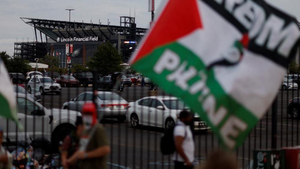 A few protestors, one waving a large flag with Palestinian colours of white, black, green and red, stand near a parking lot next to Lincoln Financial Field.