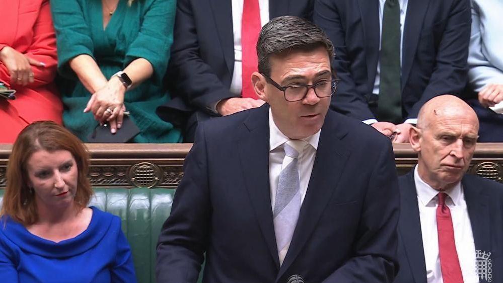 A man in a suit and with glasses and short brown hair is speaking as he stands at the despatch box in the House of Commons. A woman with shoulder length red hair and a blue top sits on the bench to his right and a man wearing a dark blue suit and red tie is seated to his left. MPs can be seen seated in the row behind.