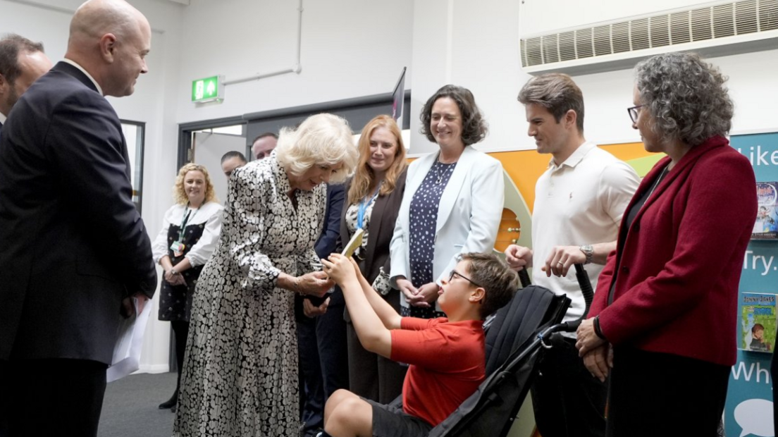 Queen Camilla in a dress of white circles on a black background is presented with a book by a child in a wheelchair wewaring a red T-shirt