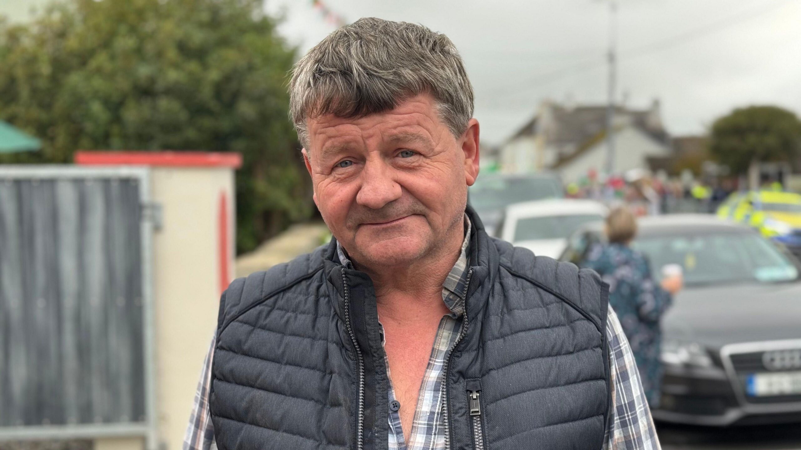 A man is standing on the street. He's looking into the camera. He has a checked shirt on with a vest jacket on. A police car is out of focus in the background.