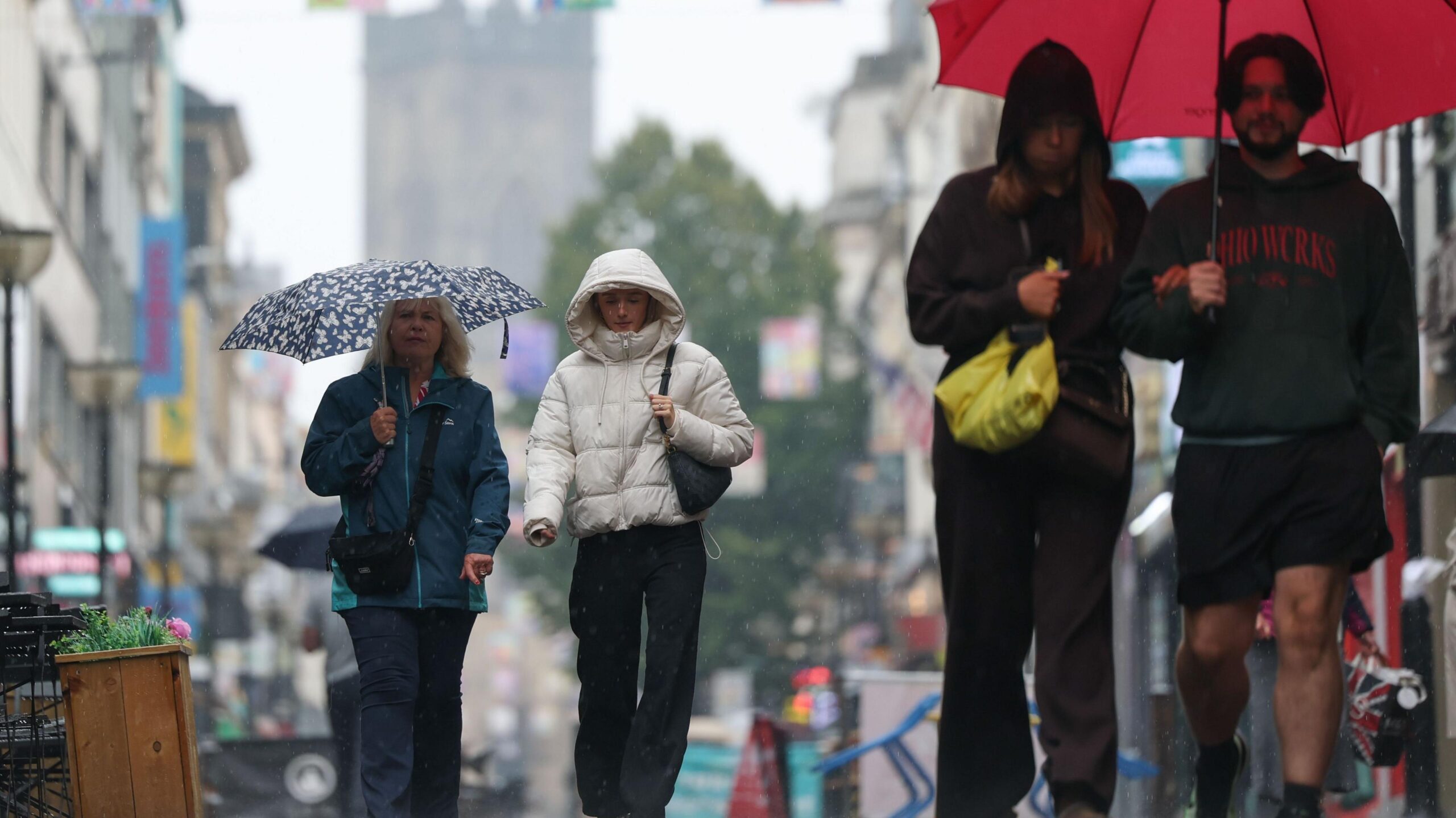 Four people, separated into two couples, walk through rain along a street with restaurants either side. They are wearing coats and each pair are sharing an umbrella.