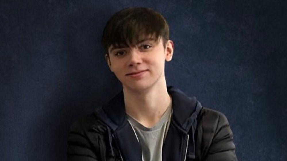 Samuel Whitaker seen on a blue backdrop wearing a grey top, dark hoodie and jacket.