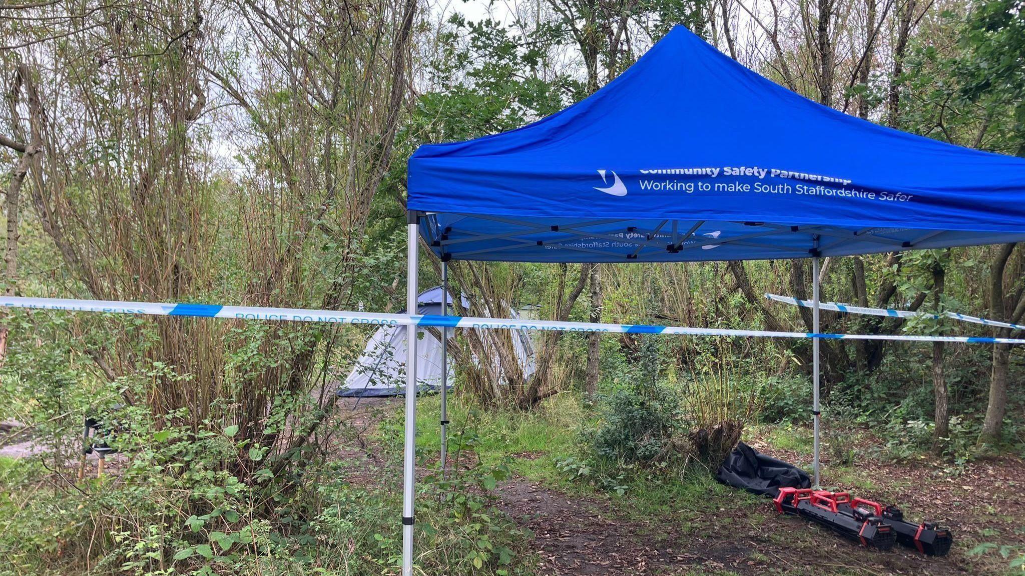 A blue gazebo with white writing on it is a woodland area. Further into the woodland is a light-coloured tent. There is blue and white police tape around the scene.