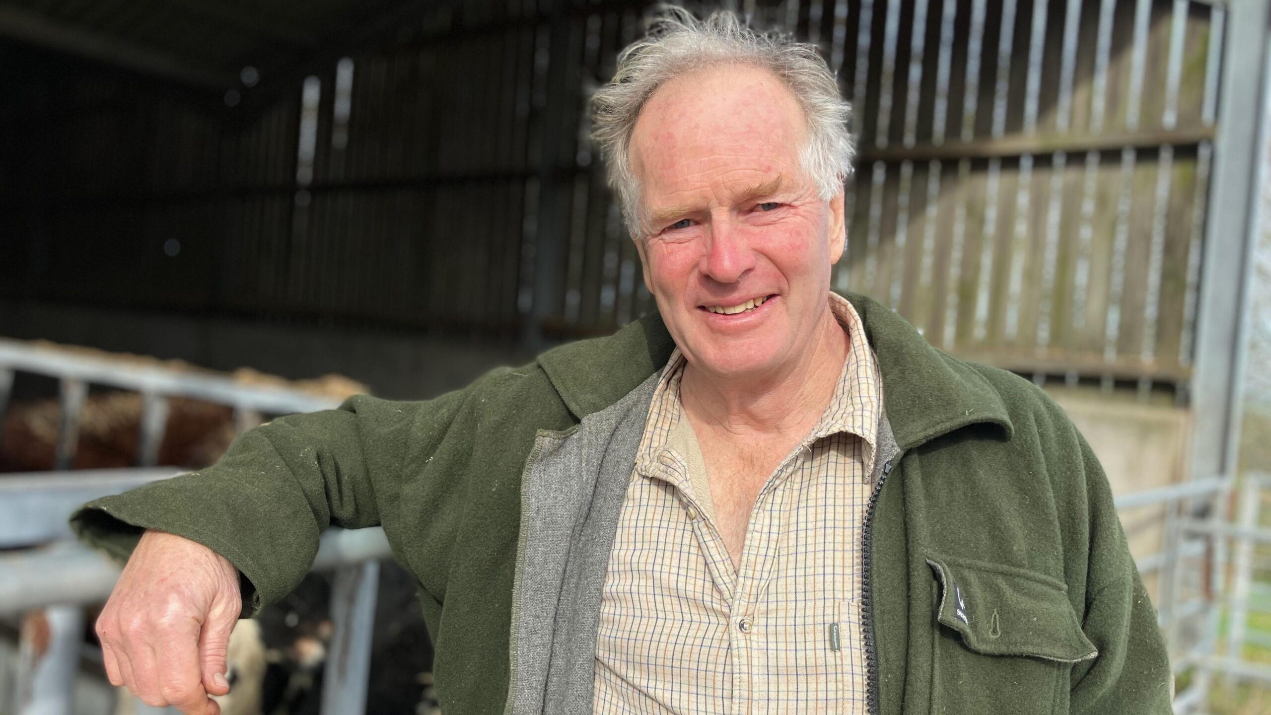 A man with white hair smiles at the camera. He is leaning on a metal farm gate. He is wearing a brown, plaid shirt and dark green jacket. In the background is an open-sided barn.