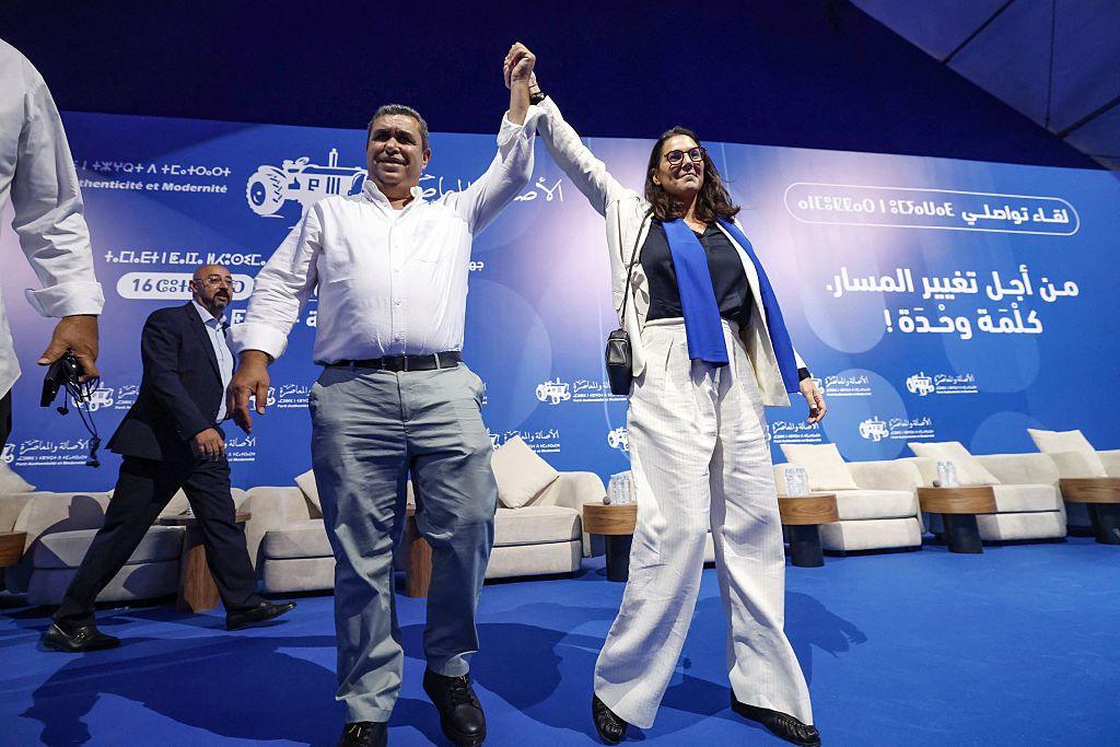 El Mansouri holding hands with a Pam colleague during one of the part's rallies. She is wearing a white suit and standing on a blue platform featuring Pam's logo in several languages.