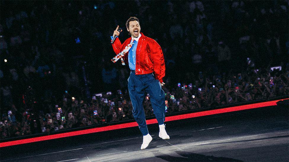 Harry Styles is wearing navy trousers with a belt and a light blue shirt with a white collar and flower patterned tie.He has a red bomber jacket over the top. He is jumping on the stage, you can see fans watching from the crowd behind him.