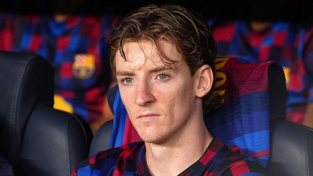 Anthony Gordon, in Barcelona's blue-and-red training kit, sits on the bench during a match