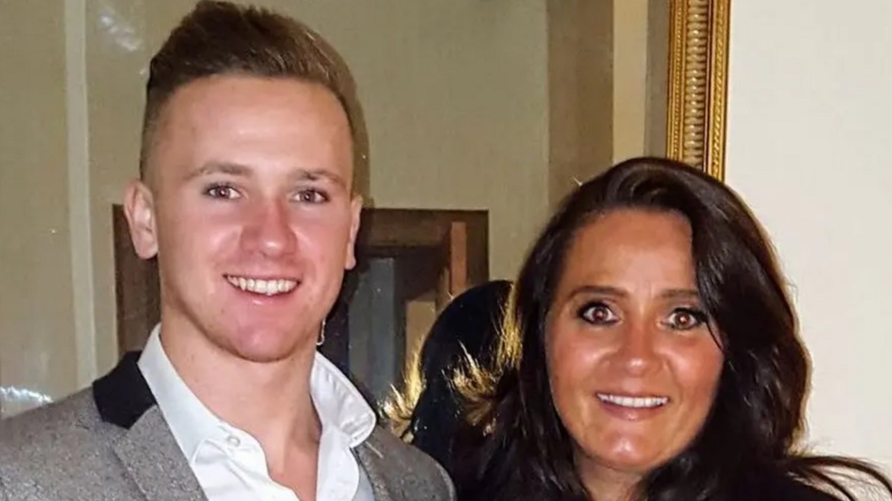 A head and shoulders image of a young man wearing a grey jacket over a white shirt and a woman. They are both standing in front of a mirror and looking into the camera while smiling.