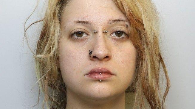 A mugshot of a young woman with blonde hair. It tied back in a messy ponytail. She has her lip, eyebrow, nose and the bridge of her nose pierced with metal bars and has a neutral expression.