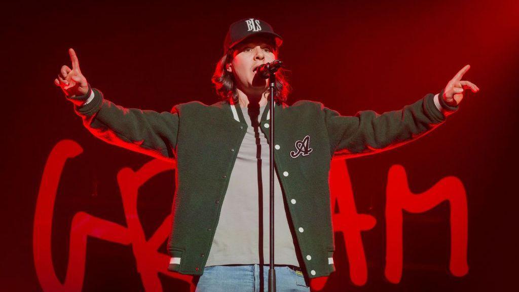 Singer of band Lukas Graham singing into a microphone on stage with both arms raised up