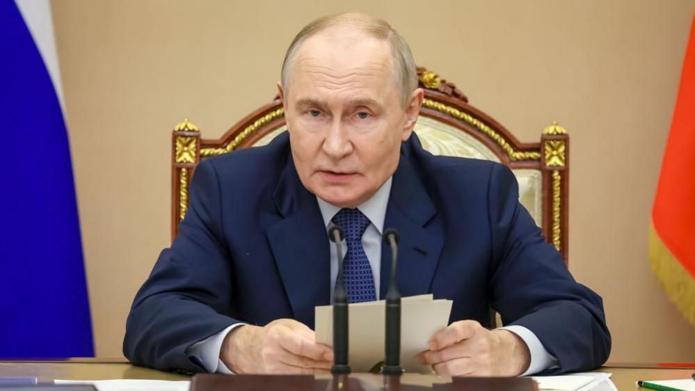 Putin sits in front of a desk and microphone holding some notes