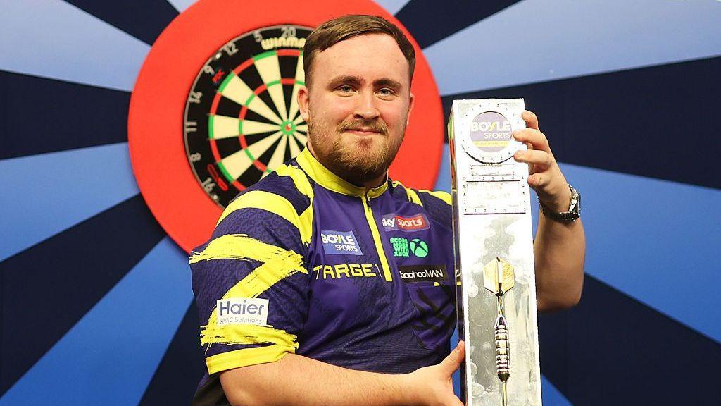 Luke Littler holding the 2025 Darts World Grand Prix trophy