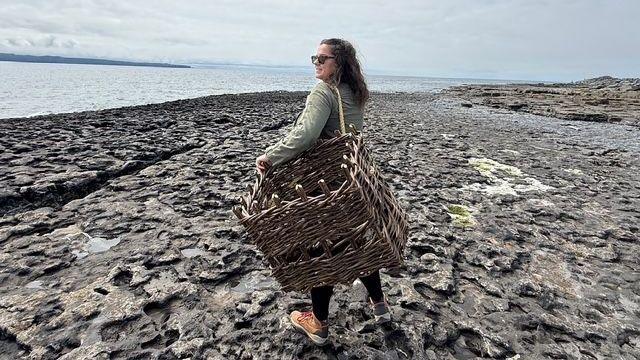 Brunette Colorado native Mary Timby, 39,  wears sunglasses and tennis shoes as she holds a woven basket over her shoulder on an Irish shoreline, the sea and broody skies visible in the background