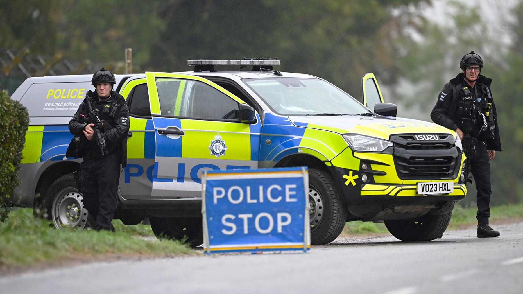 Two armed police men at Fairford standing next to a yellow police van with a blue sign saying 'police stop' on the road in front of them