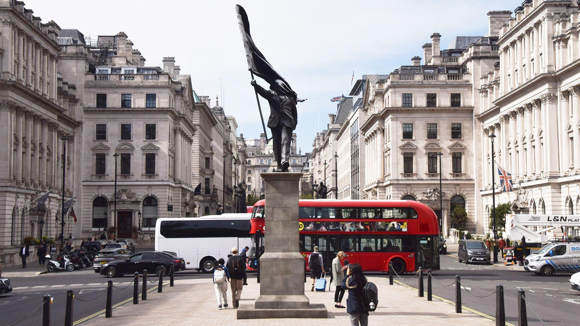 A grey statue of a man wearing a suit, walking forward off a plinth, carrying a flag that covers his face. A red London bus can be seen in th bcakground