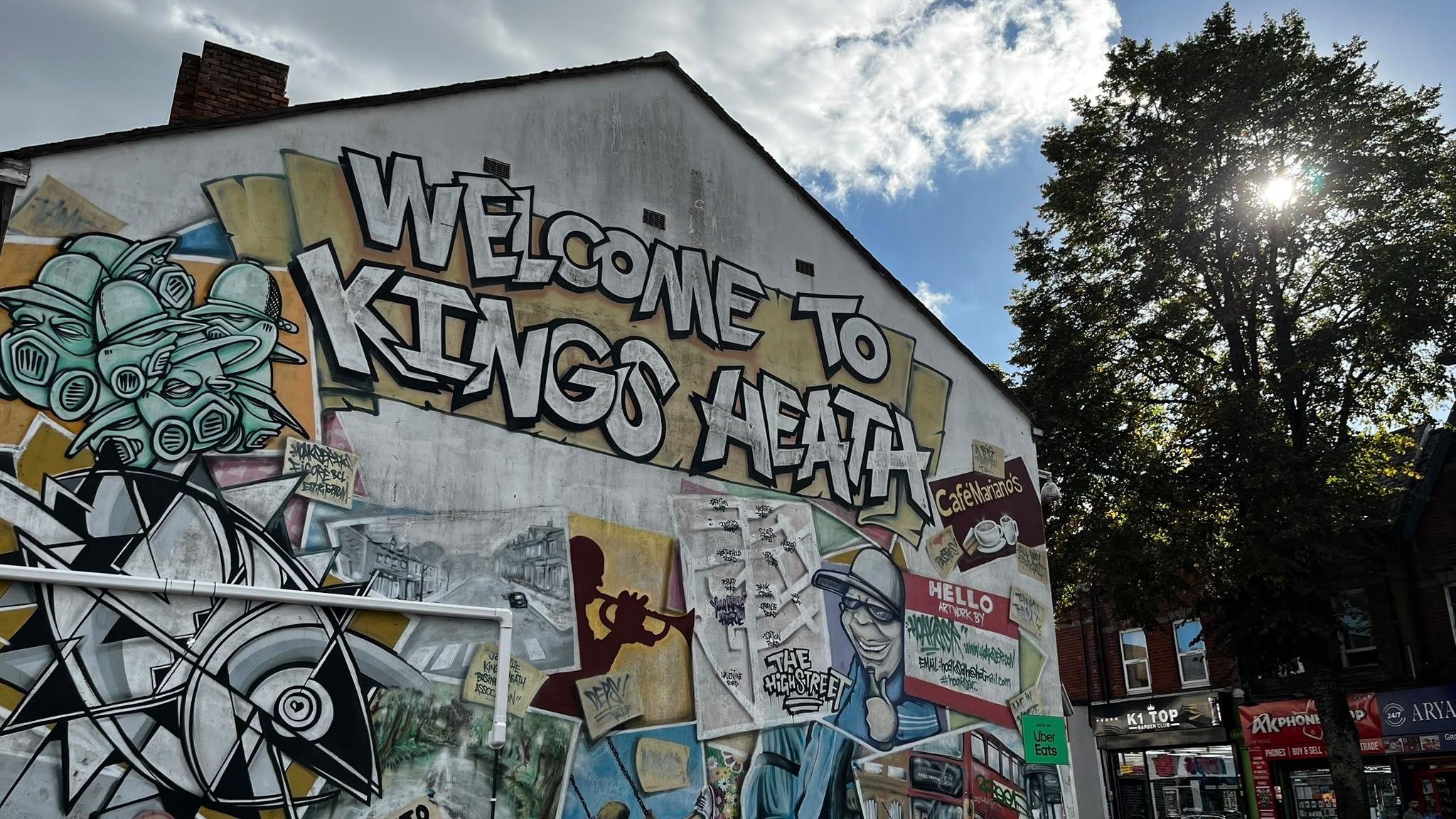 A large mural on the side of a white building which reads: Welcome to Kings Heath. To the right is a large tree and the sun is shining through the branches. Blue sky is visible with cloud over most of the top of the image.