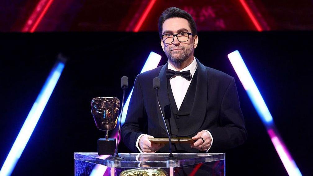 A man wearing glasses and a three-piece suit with bow tie stands on-stage behind a lectern, holding an envelope in both hands. On the lectern are two microphones and a Bafta award - a golden trophy shaped like a theatrical mask.