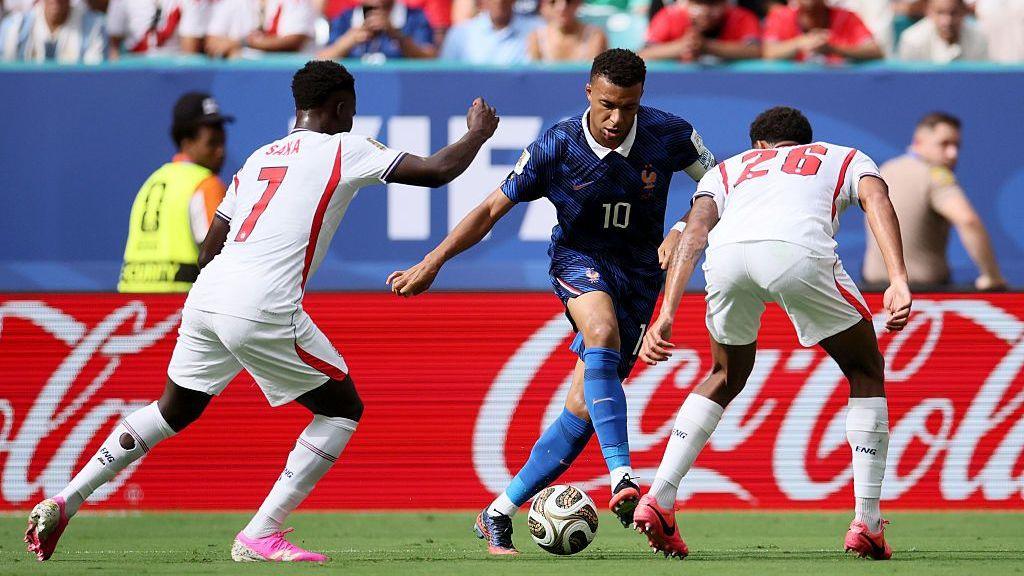 Kylian Mbappe dribbles with the ball while Jarrell Quansah and Bukayo Saka close him down during the 2026 FIFA World Cup third-place play-off match between France and England at the Hard Rock Stadium in Miami, Florida. In the background an advertising hoarding bearing the Coca Cola logo is visible