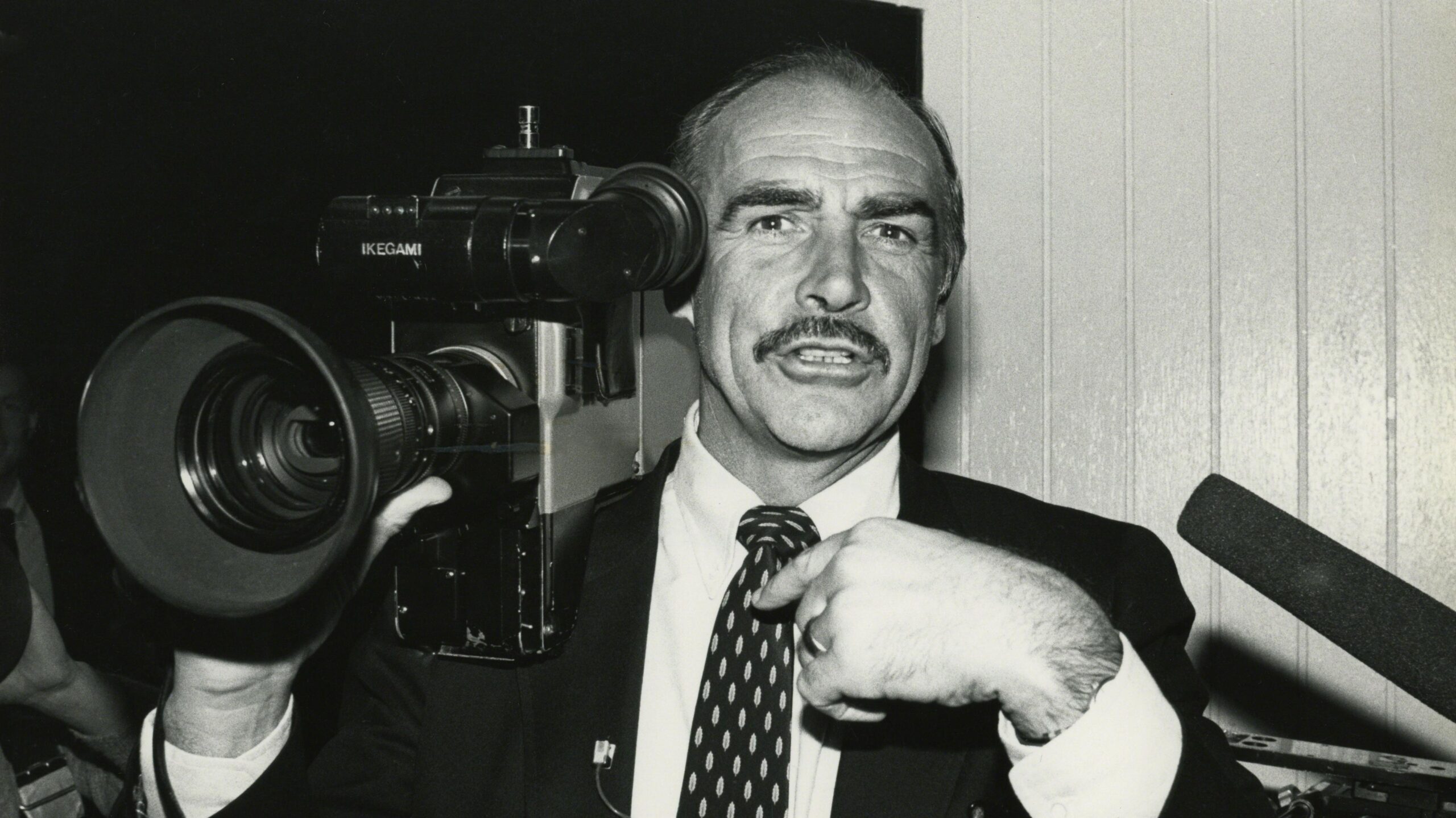 A black and white photo of a man holding an old television camera wearing a suit and tie.
