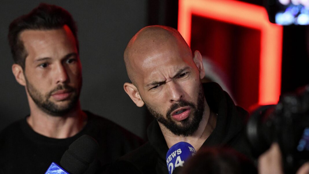 Tristan and Andrew Tate delivering a press statement. The brothers are dressed in black and Andrew has a disgruntled facial expression as he speaks into a microphone.