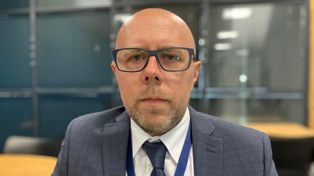 A head and shoulders shot of Det Sgt Rich Glanville wearing a grey suit and white collared shirt with a navy tie. He is bald, with light stubble and black rectangular glasses.