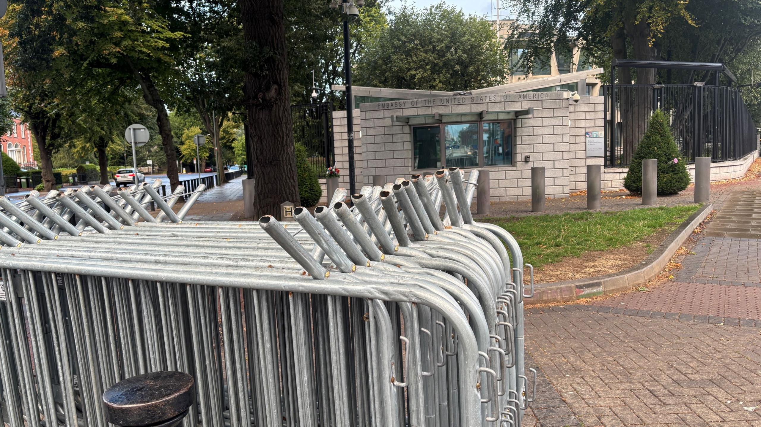 Metal crowd control barriers on footpath outside American embassy in Dublin. Trees are visible beside in front of the embassy. The pavement is red bricks and grey bricks.