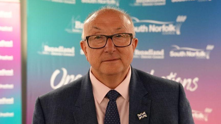 Colin Sutton standing in front of a council board. He is wearing a suit with a grey blazer, a light pink shirt and a patterned tie. He is looking directly at the camera and smiling.