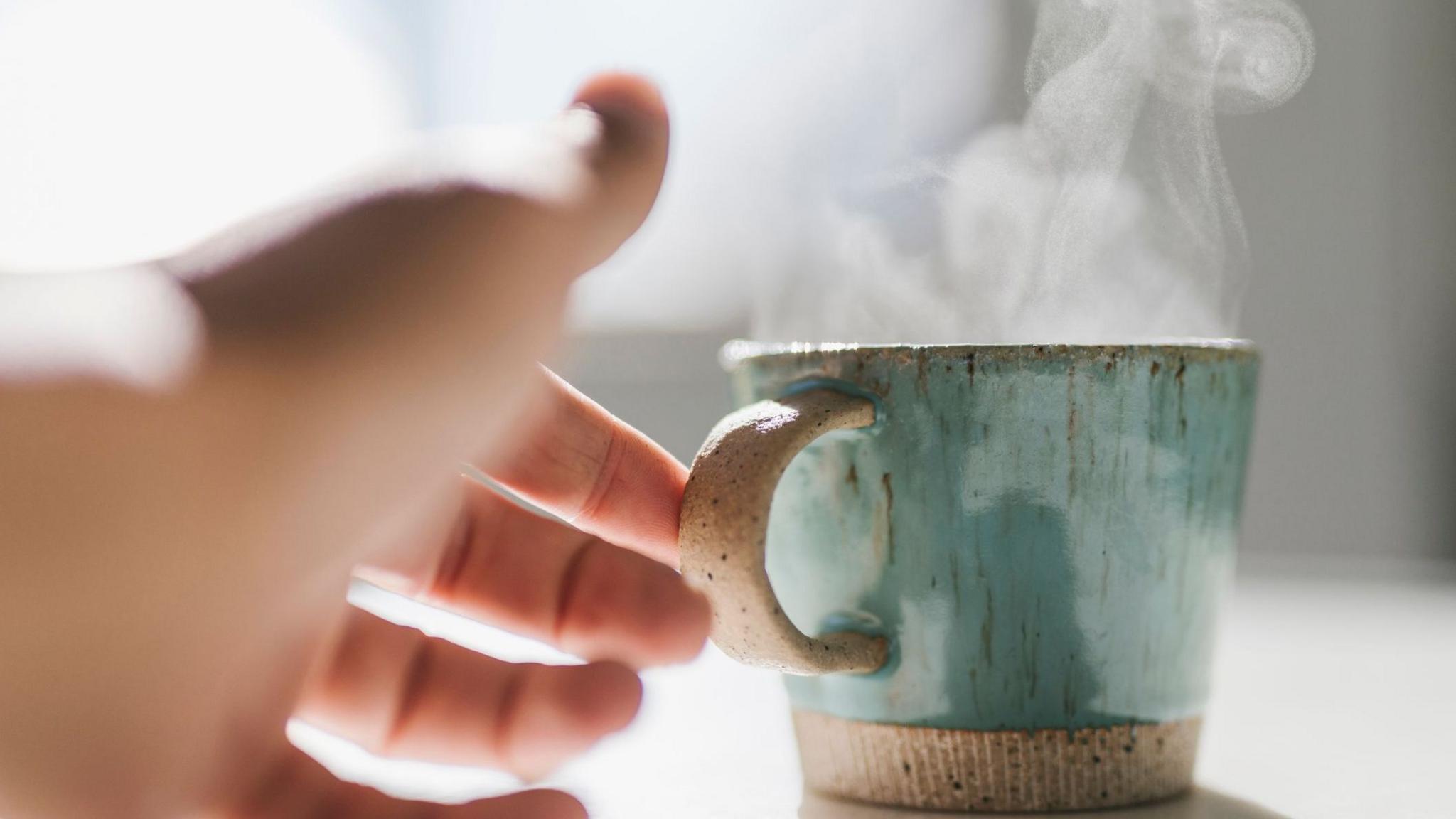 A woman reaches her hand out to pick up a steaming hot mug which is shiny and green-coloured