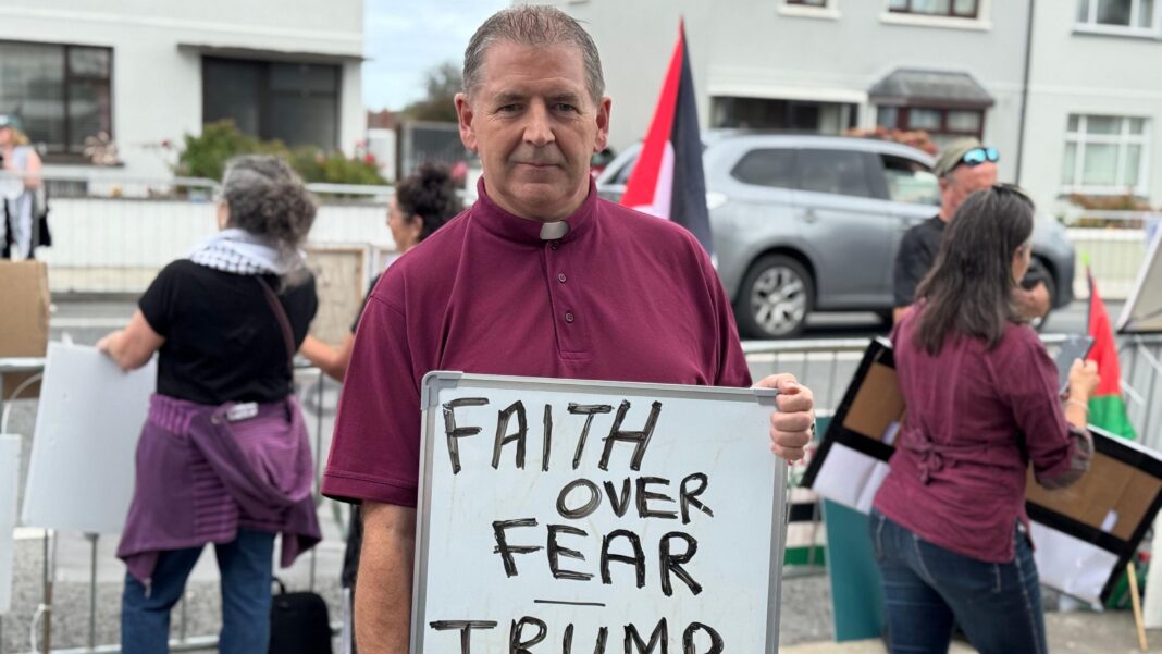 Trump doubles down on united Ireland comments on day two of visit A man wearing a white collar on his maroon top is holding a white board saying faith over fear. Trump get out of here.