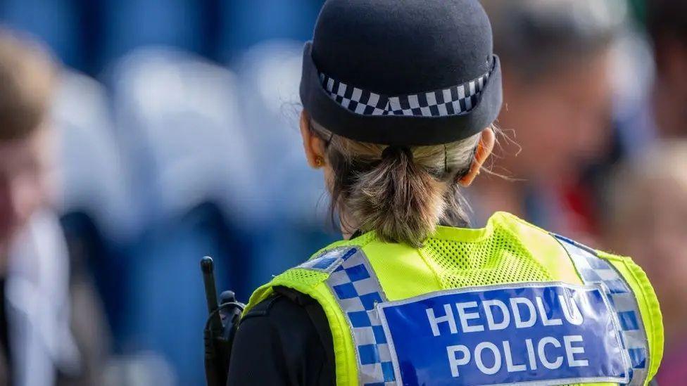 A female police officer wearing a hat and uniform faces away from the camera. Her uniform reads: HEDDLU POLICE