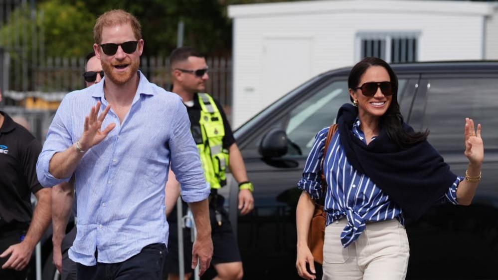 Harry and Meghan waving at a crowd, several security staff and a black car are behind them on 17 April 2026