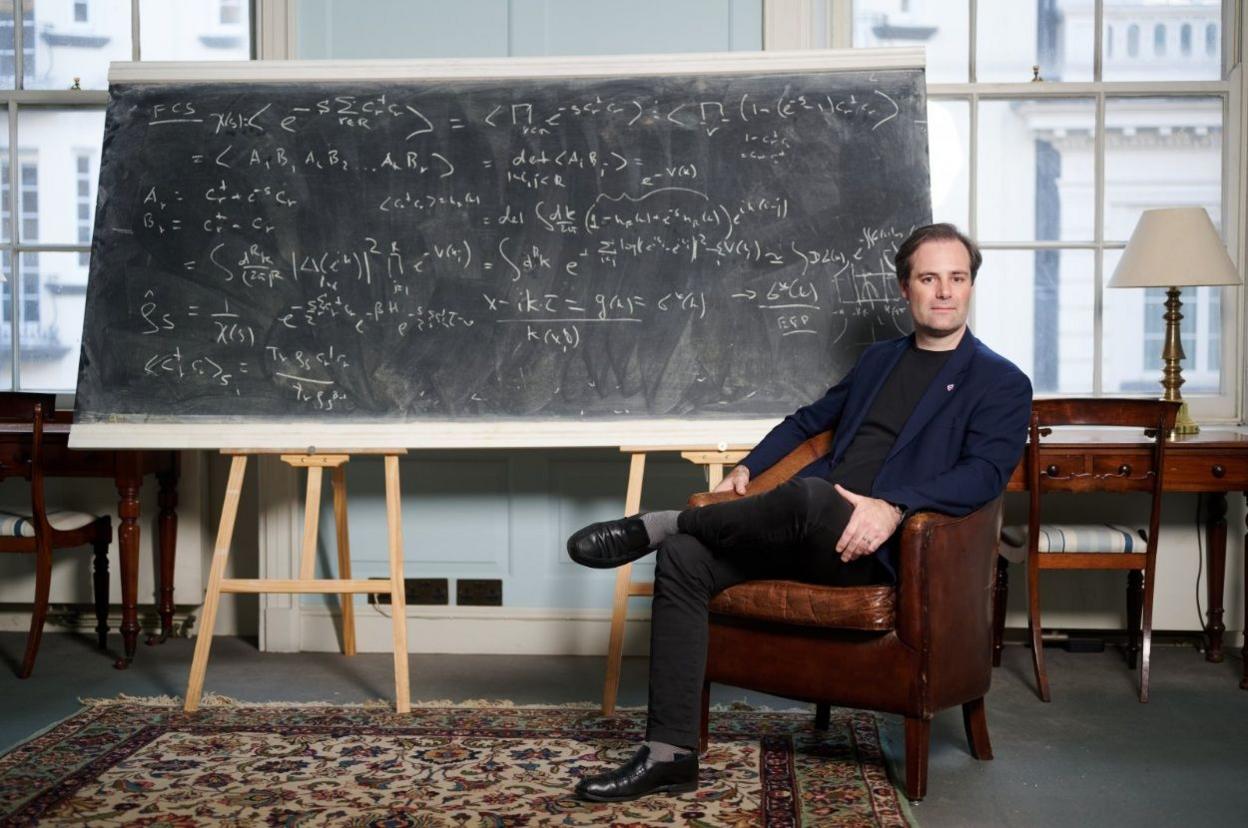Ben Delo sitting on a leather armchair with his legs crossed, wearing a blue jacked and black trousers, sitting in front of a blackboard with mathematical equations written in chalk.