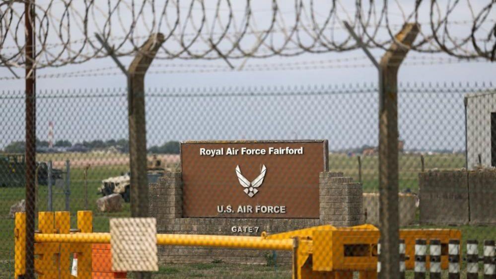 A sign stands behind a fence at the RAF Fairford airbase. Two vehicles can be seen in the background on a field behind the sign