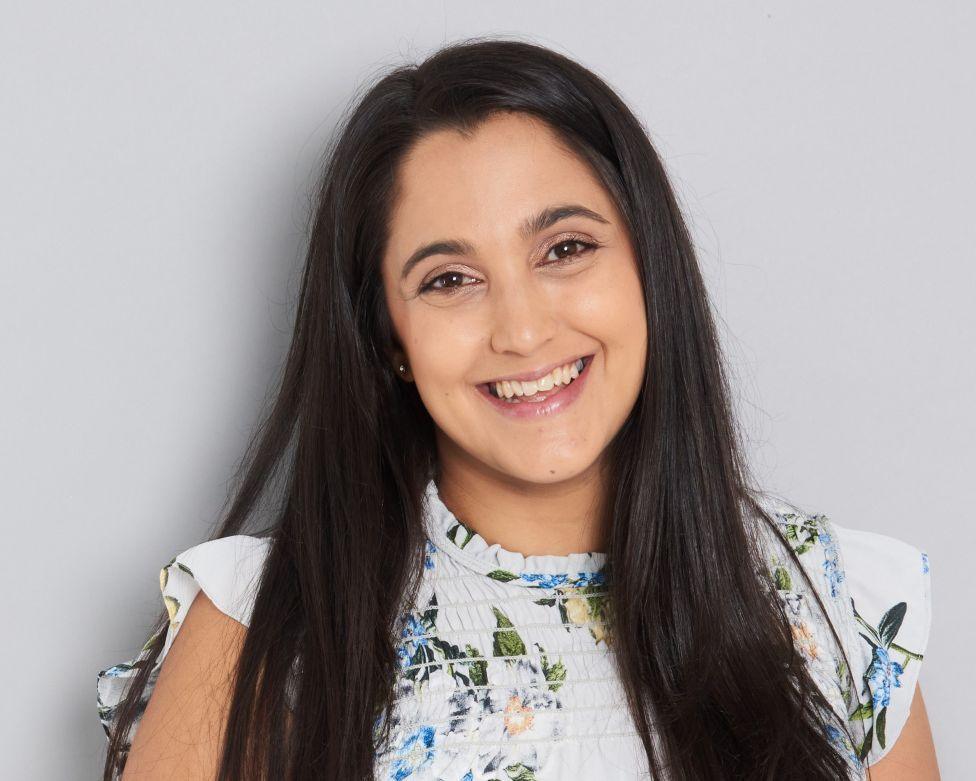 A woman in a white floral top stands in front of a plain grey wall and smiles