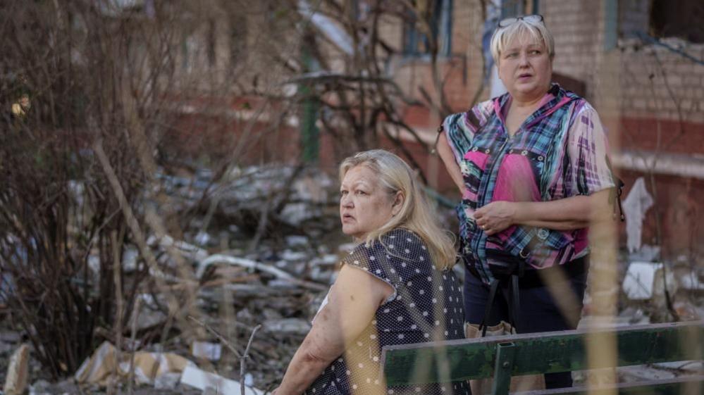 Two middle-aged women stare into the distance amid the rubble from a Russian attack
