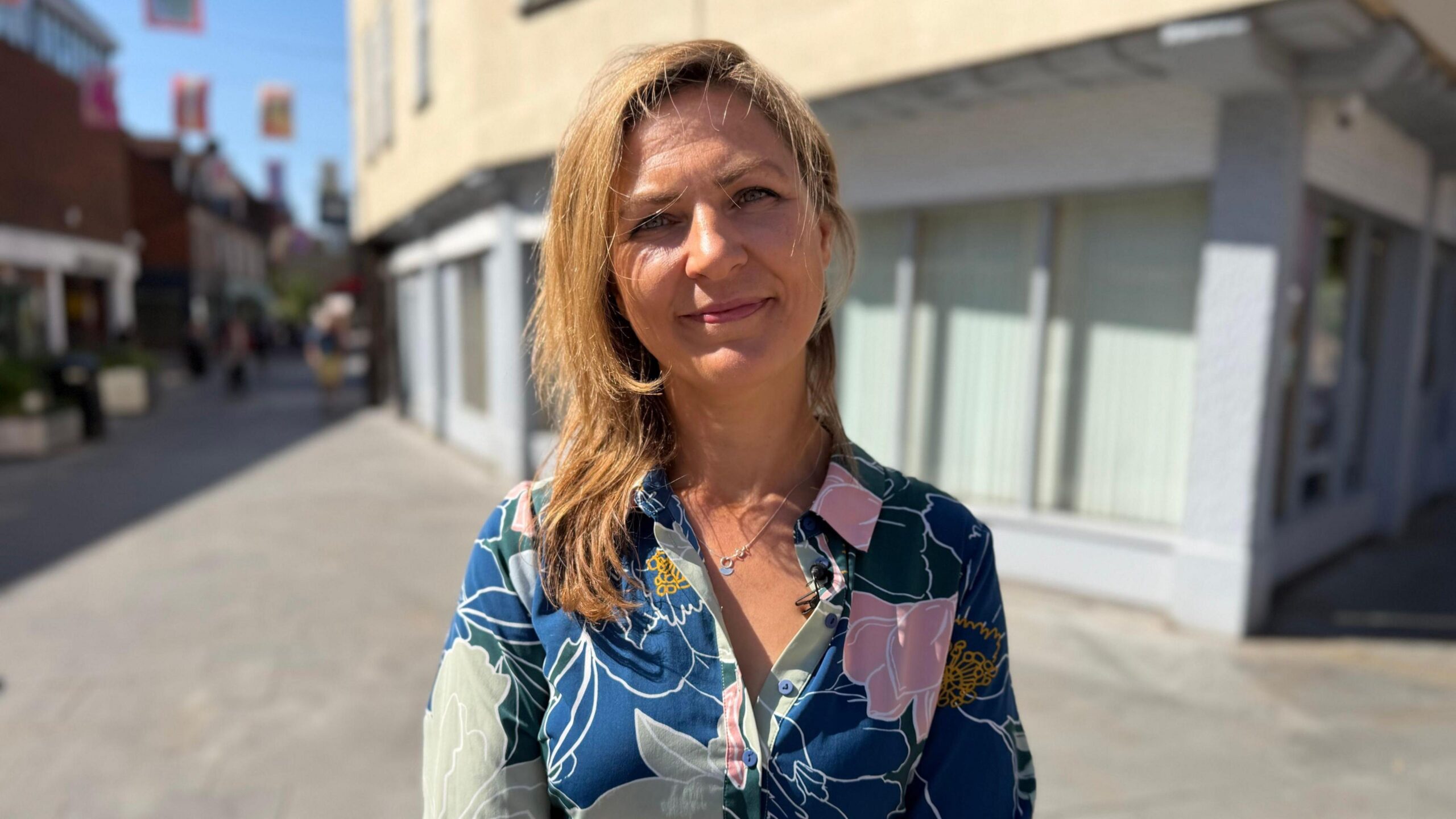 Woman stands on the high street. She has light brown hair and is weating a blue floral shirt.