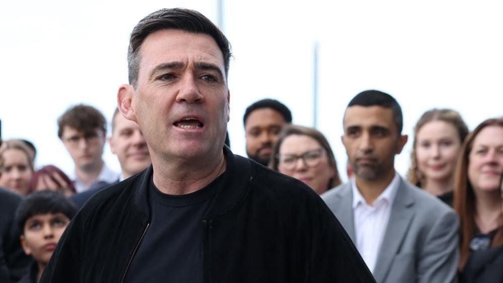 Andy Burnham is wearing a dark jacket over a dark top. He is stood in front of a crowd of people and is speaking.