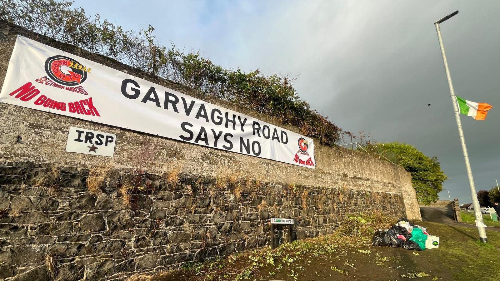 A sign pinned to a wall saying Garvaghy Road say no. It's cloudy day. At the end of the wall in a lamppost with an Ireland tricolour flying from it.