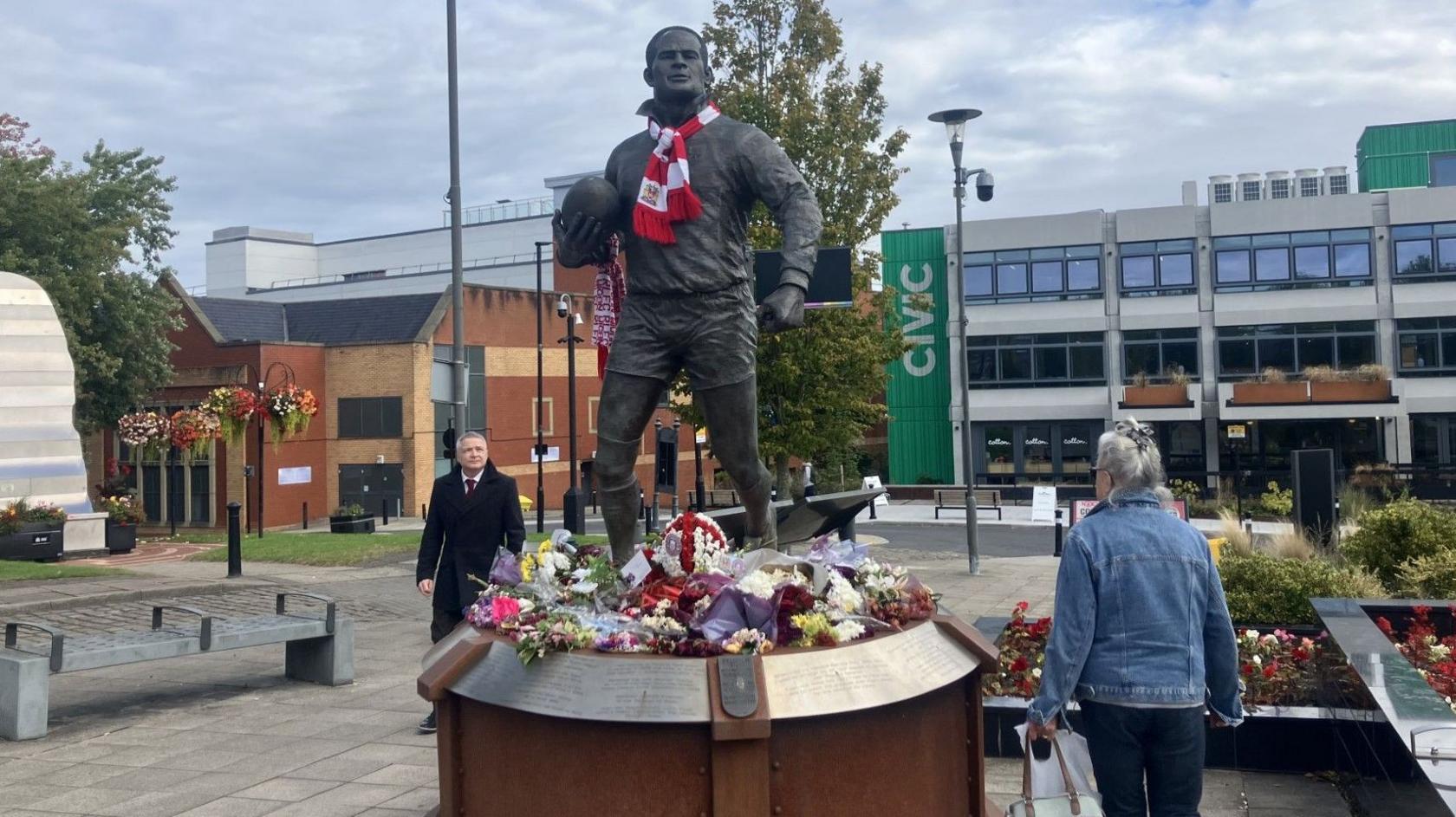 A statue of Billy Boston holding a rugby ball. He had a red and white scarf around his neck. Flowers surround the base.