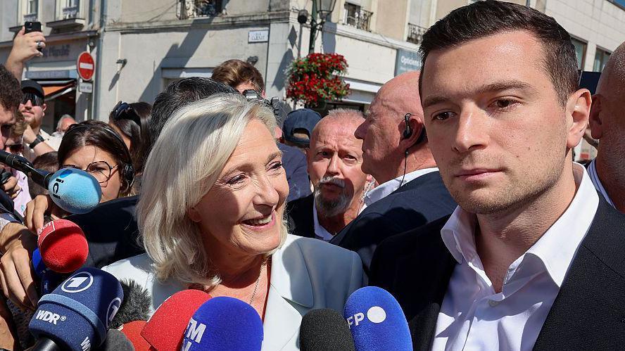 A woman stands in a crowd smiling in front of microphones while a man to her right stands beside her with no expression