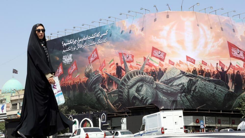 A woman clad in black walks in front of a poster that shows a toppled Statue of Liberty in front of a crowd waving red flags