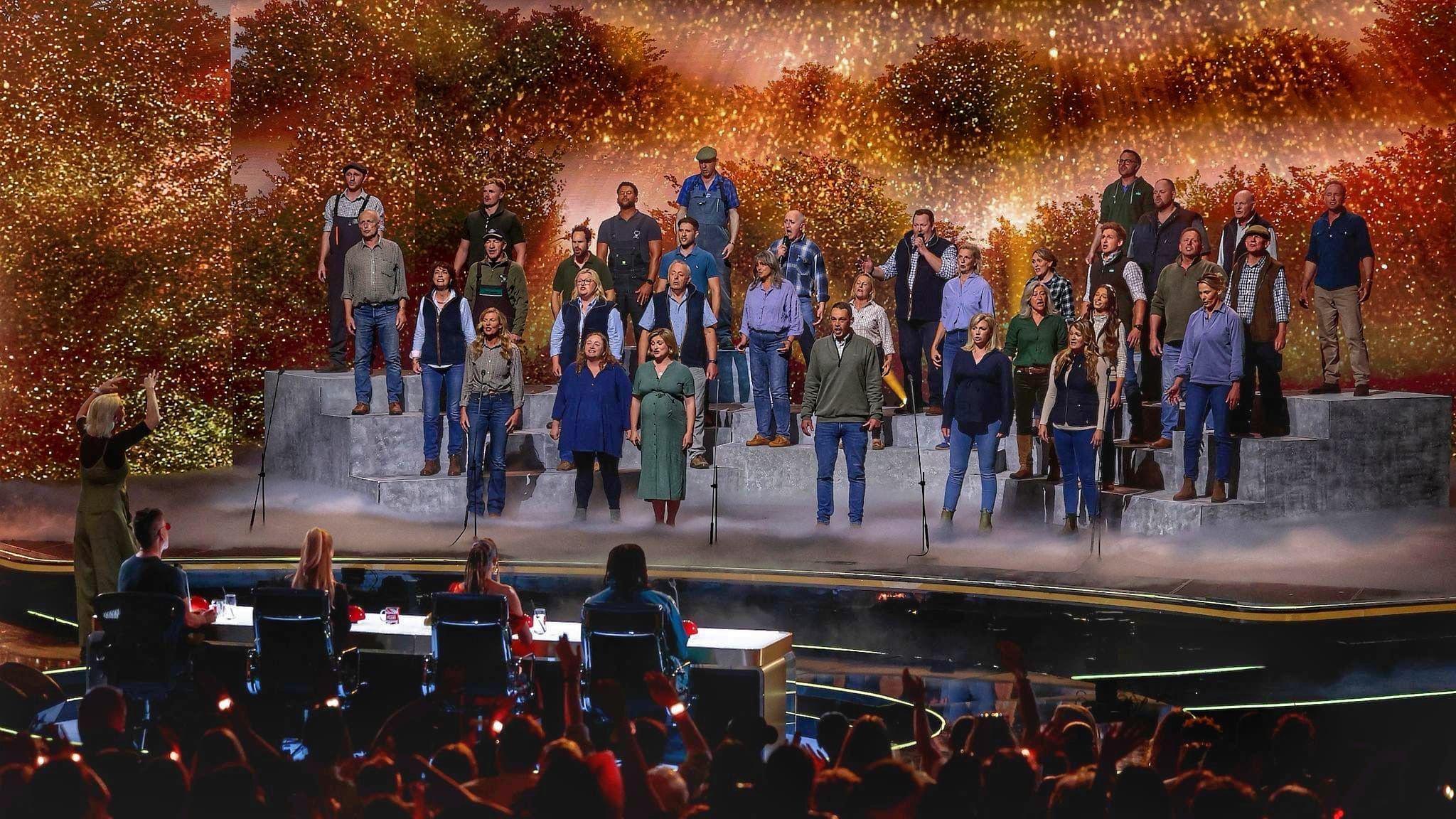 A large group of performers stands on tiered platforms on a brightly lit stage, appearing to sing together as a choir. A conductor stands at the front, guiding the performance. In front of the stage, a panel of judges sits at a table, watching. The background features warm, glowing lights and foliage-like scenery, while an audience in the foreground raises hands and watches the show.