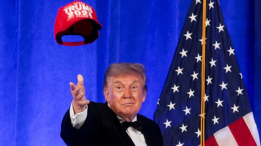 Trump in bow tie, before American flag, tosses red 