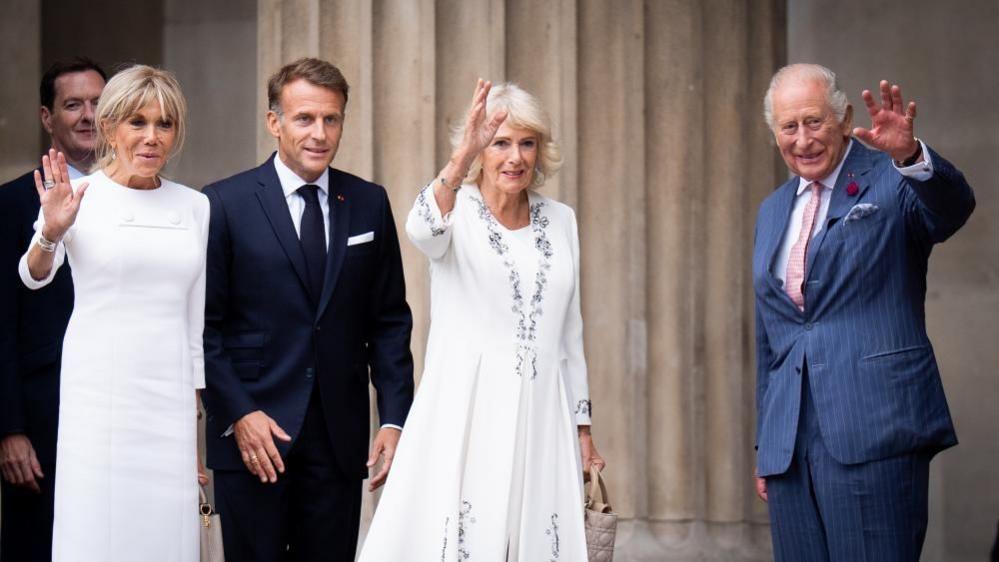 Brigitte and Emmanuel Macron stand next to Queen Camilla and King Charles. The two men are wearing blue suits while both women are wearing white dresses. They are waving to the camera outside the British Museum.
