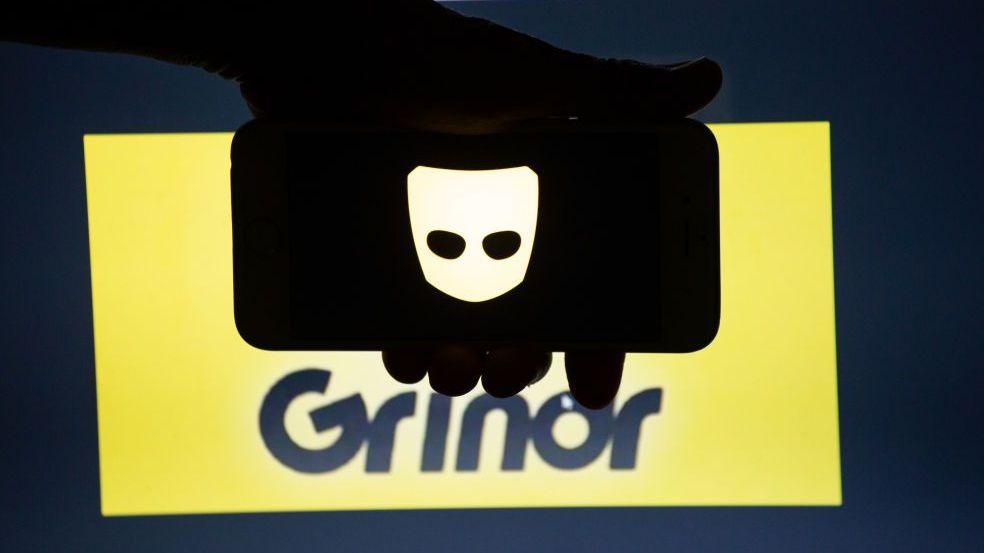 Grindr's app logo, displayed on a smartphone screen, is held in front of a yellow and black backdrop bearing the company's logotype.