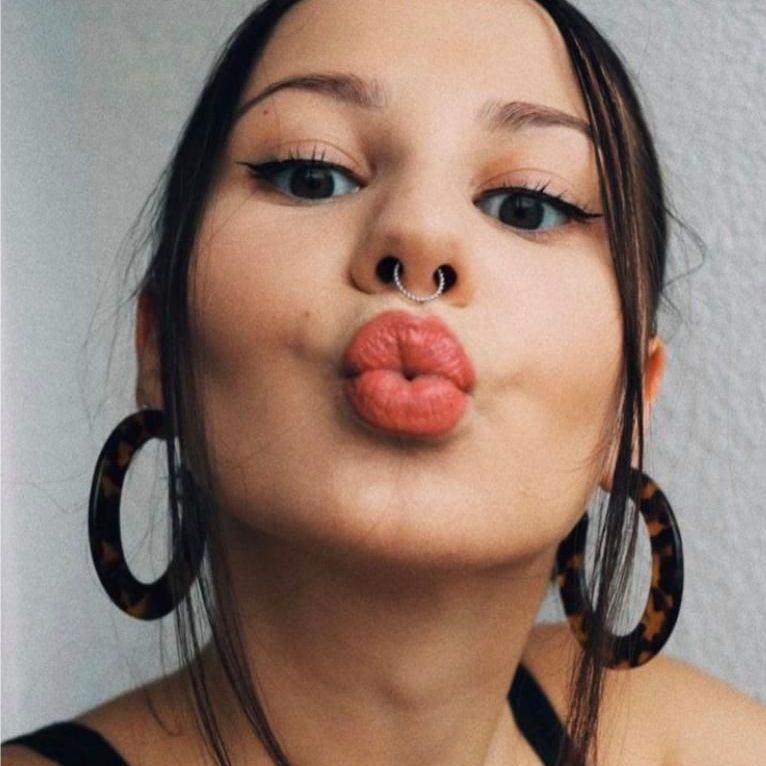 A young woman with brown hair and wearing bright red lipstick pouts at the camera. She is wearing a black vest top.