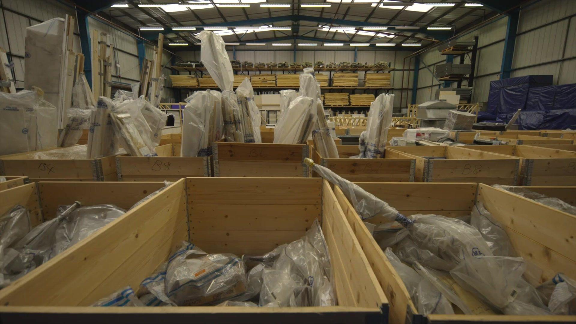 A large warehouse filled with wooden crates containing items wrapped and sealed in clear plastic, with stacks of materials stored on shelves in the background.