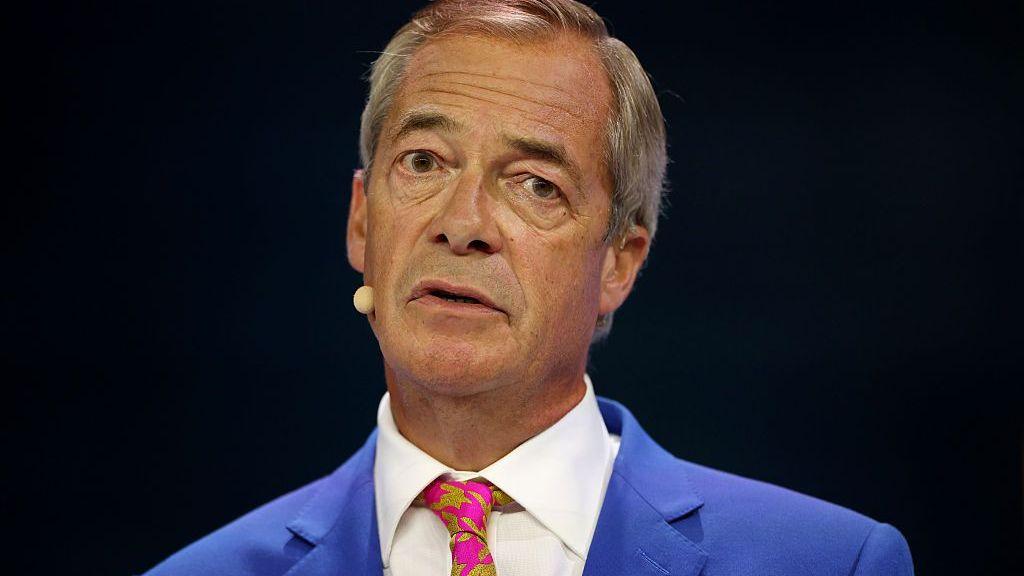 Reform UK Party Leader Nigel Farage speaks on stage. He wears a bright blue suit jacket, white shirt and a pink and green patterned tie. He is stood in front of a black background.