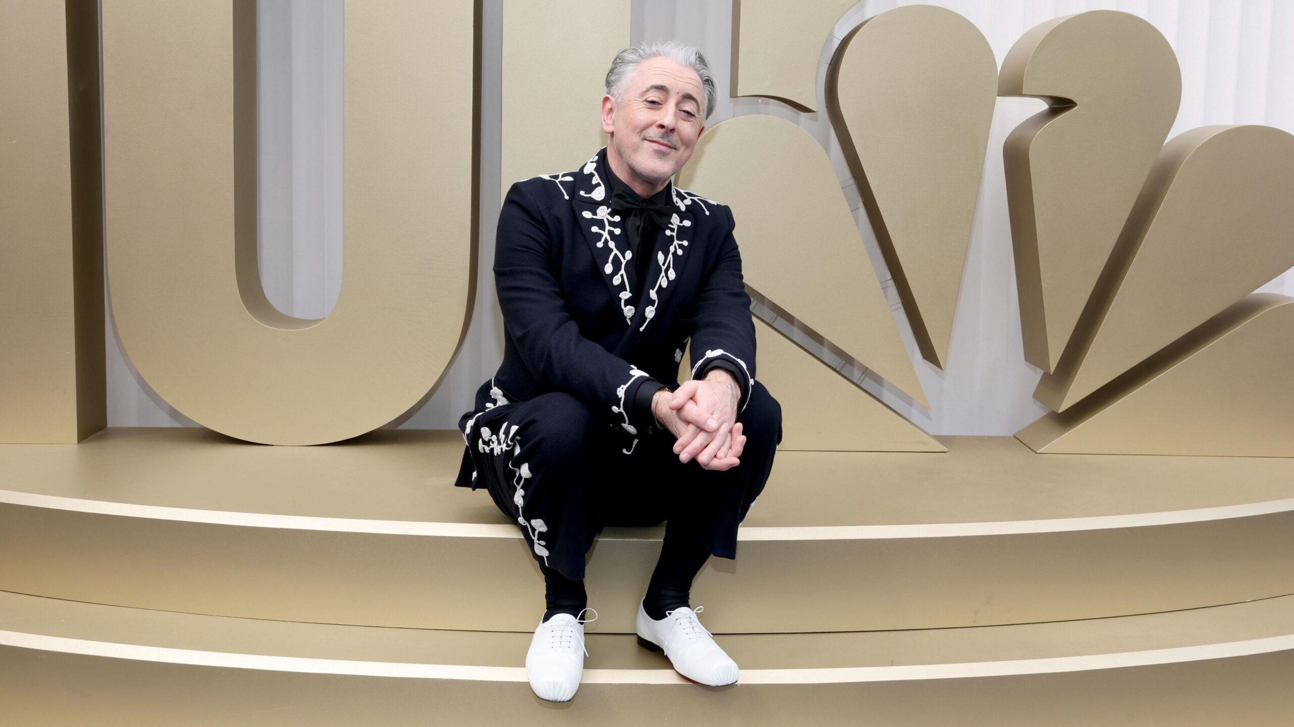 Alan Cumming in a dark blue suit with white floral embellishments, sitting on a step on the red carpet