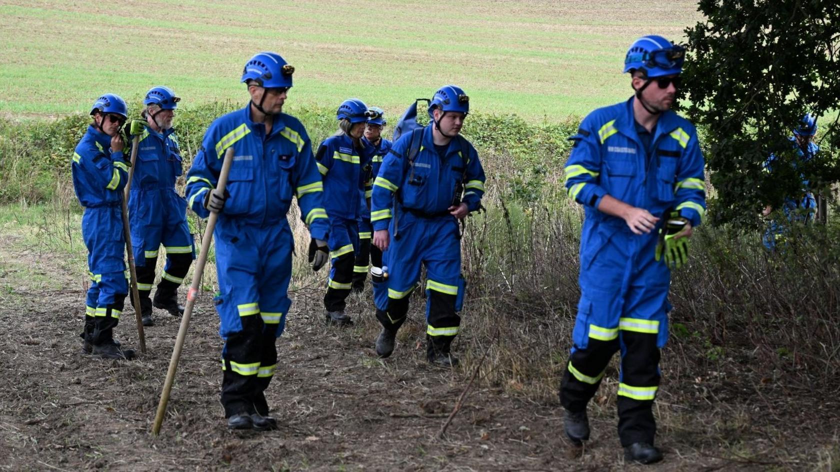 Seven people - all dressed in blue overalls with blue helmets on - searching a field for Noah Woods.